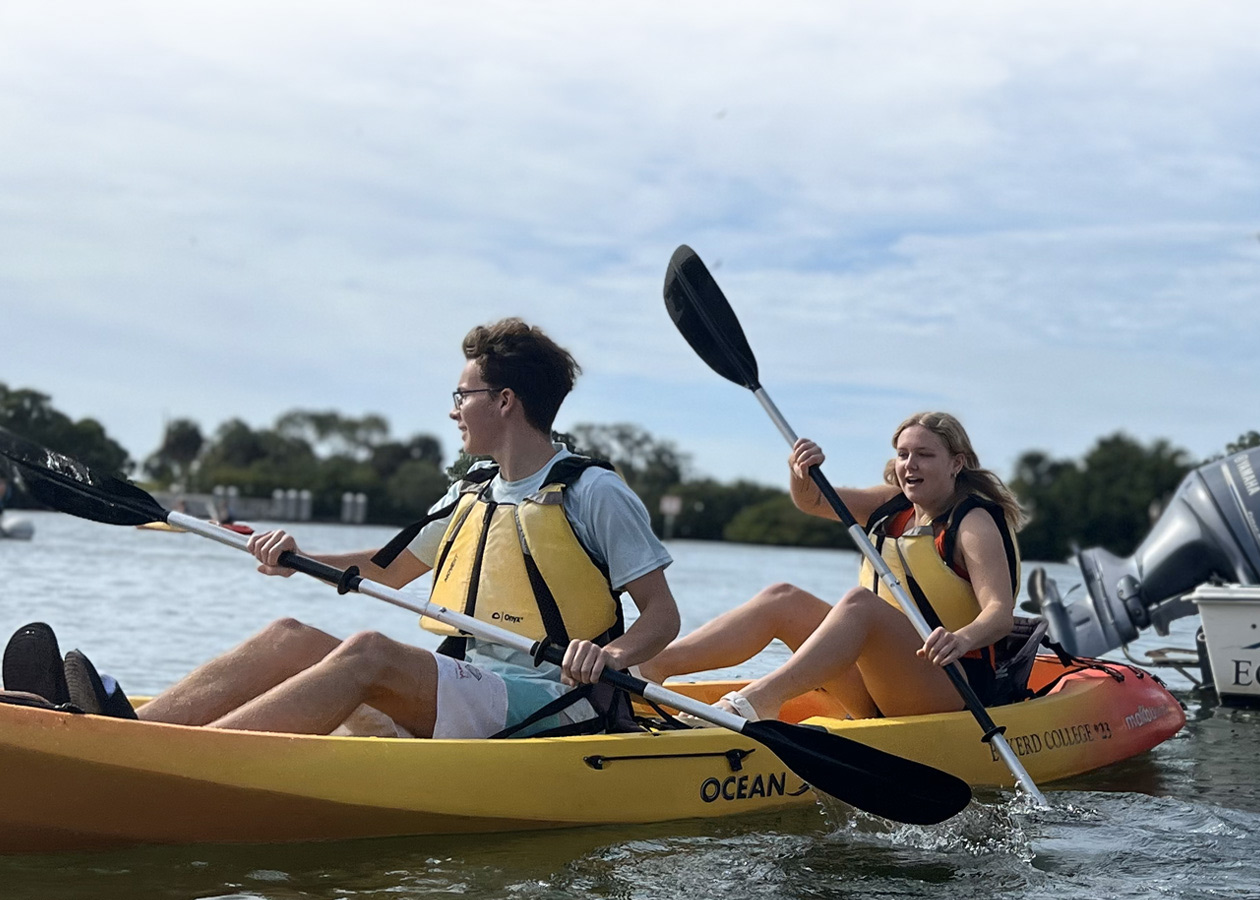 Two students wearing vests launch kayaks from a dock