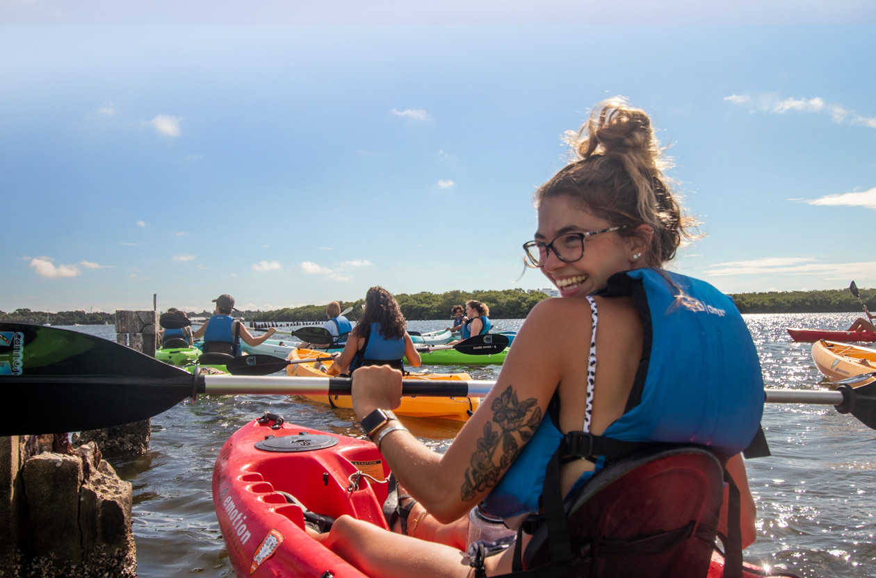 College student with tattoo wearing glasses in a kayak on the water smiling at camera