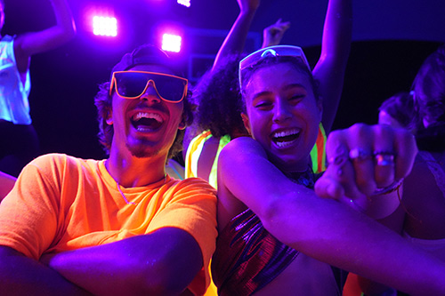 Two students in sunglasses and neon clothing smiling while dancing in the night