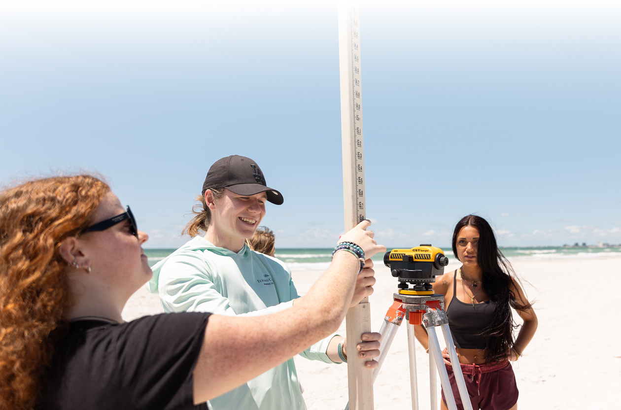 Three students standing on a beach while using a tall measuring stick and device on a tripod