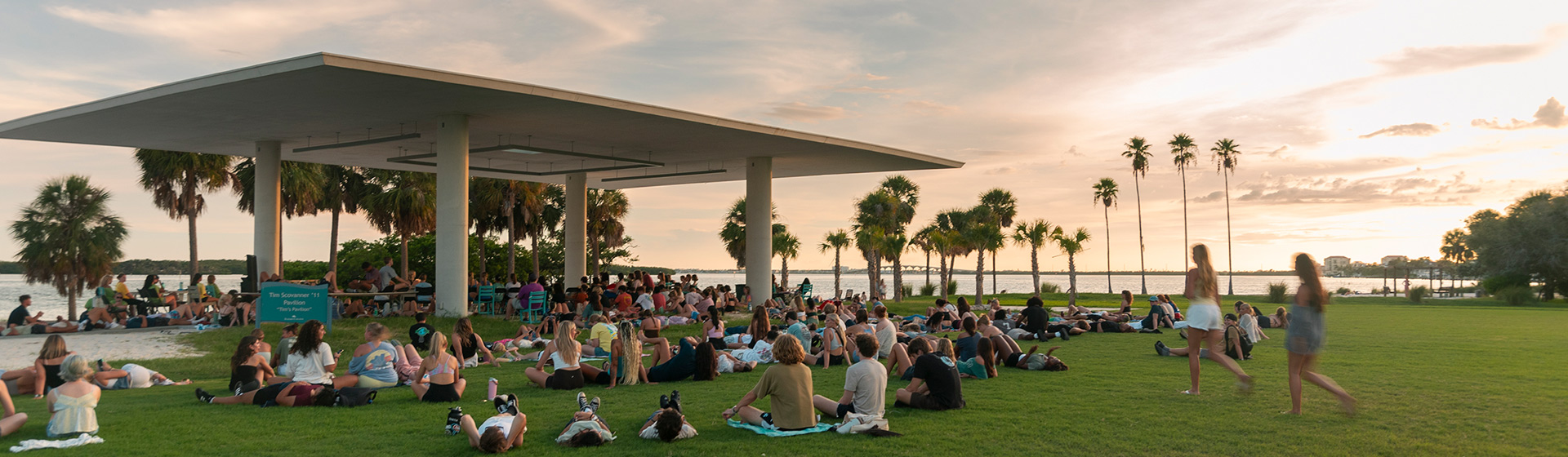 Students sit under and around am outdoor pavilion surrounded by palm trees at sunset