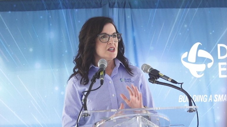 Woman speaking in front of two standup microphones with "Duke Energy" on sign behind her