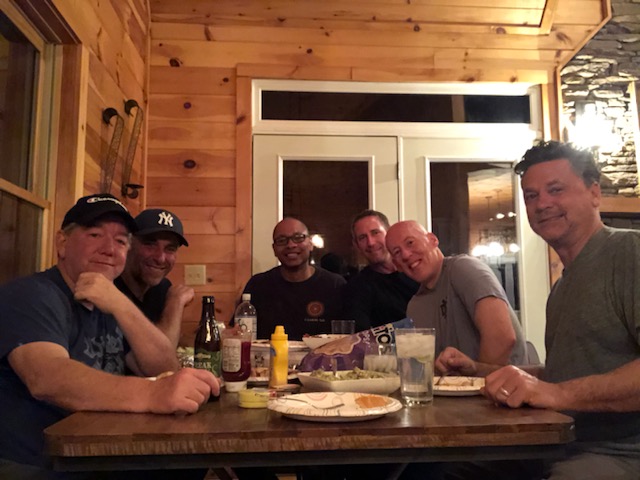 Six men seated around a wooden table while sharing a meal and smiling