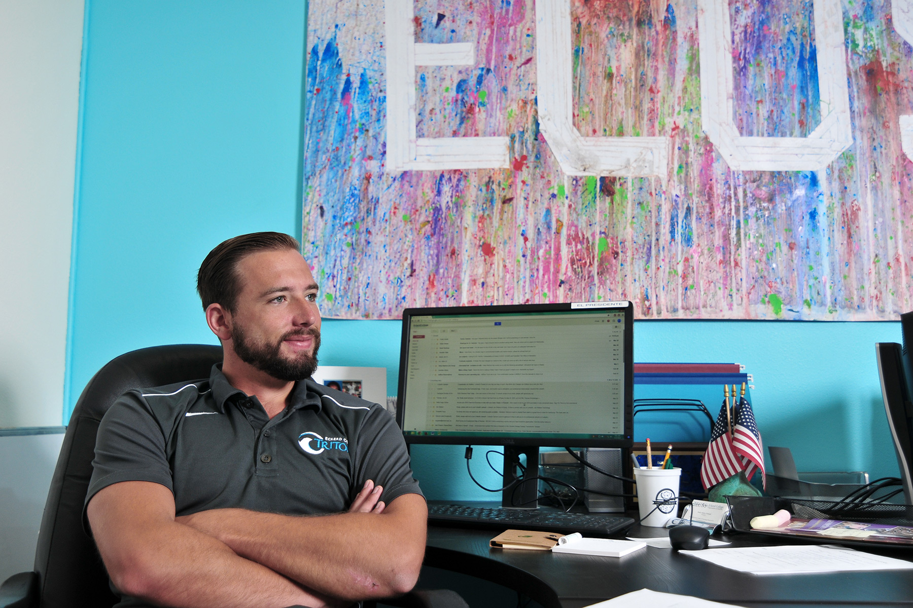Student with beard and arms crossed sits at desk with sign that reads ECOS