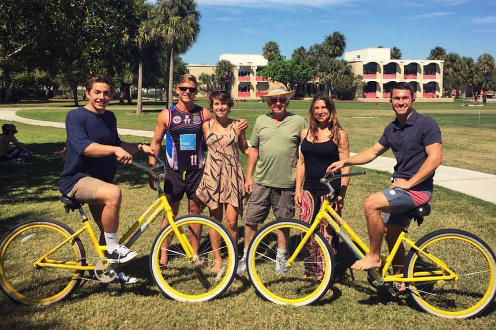 Six people standing side by side with two yellow bikes on a college quadrangle