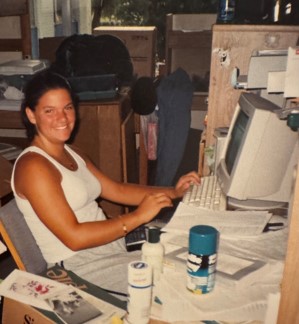Vintage photo of a student in a white tank top smiling while working at an old desktop computer with papers all over the table