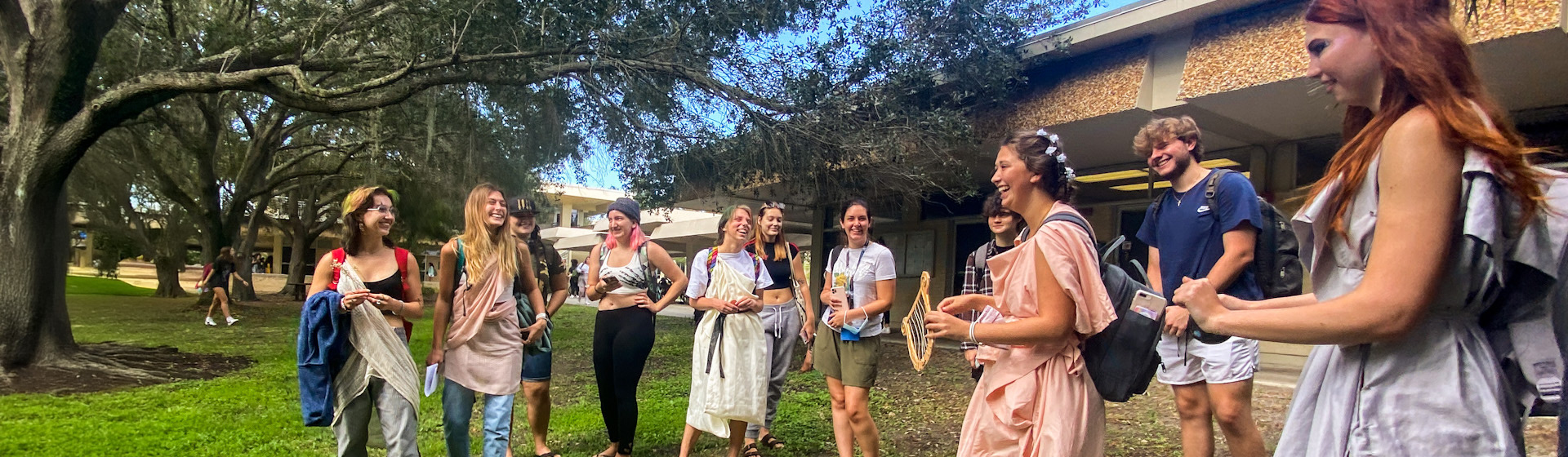 Students dressed as muses and other characters from ancient history stand in a circle on a college quad