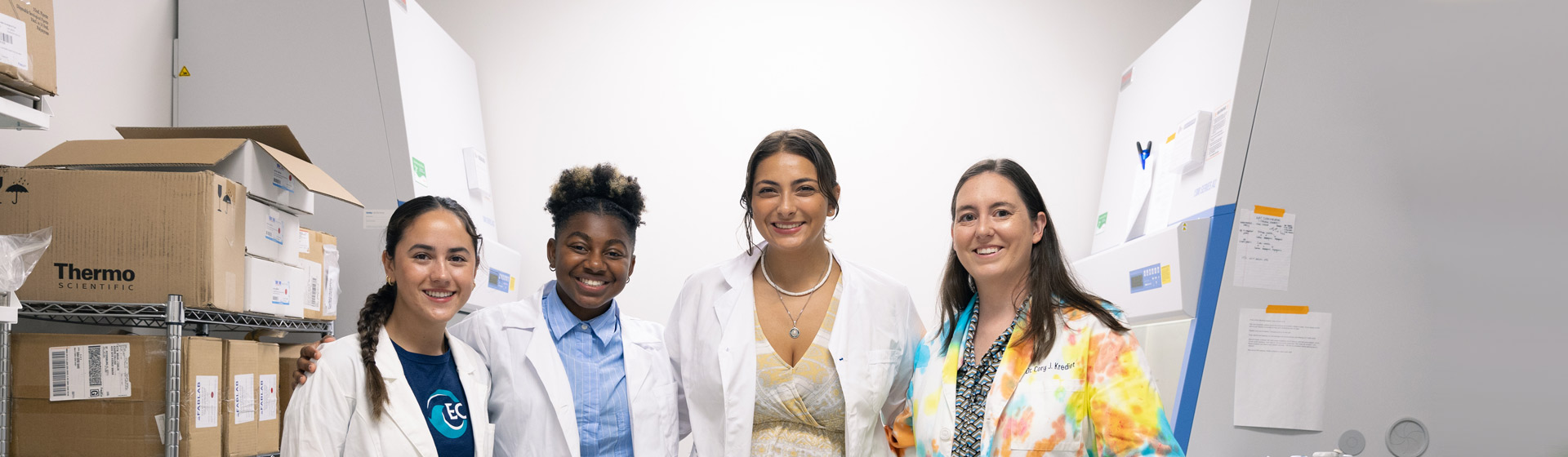 Professor and three students stand in a lab wearing lab coats