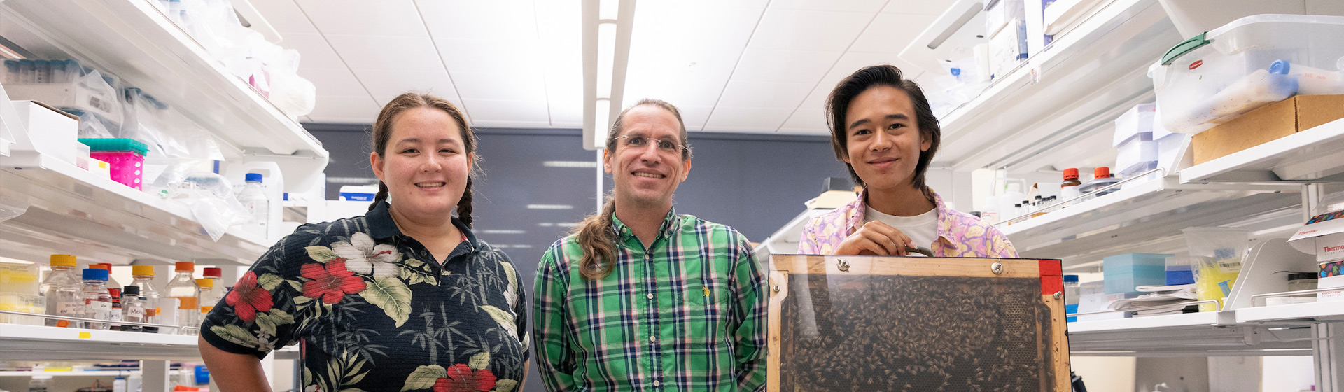 Professor stands side by side two students in a laboratory, one holding a slat full of honeybees