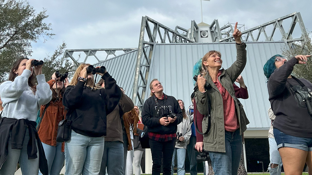 Professor stands among group of students wearing binoculars and pointing to the sky