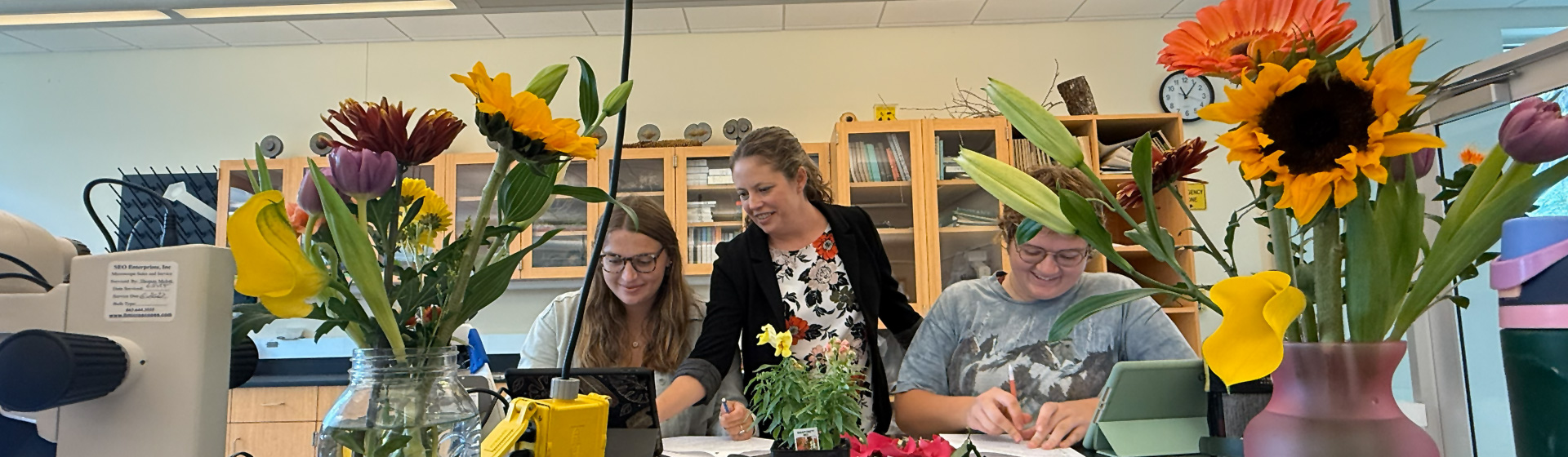 Two students seated at a lab table covered with flowers look at plants with a professor