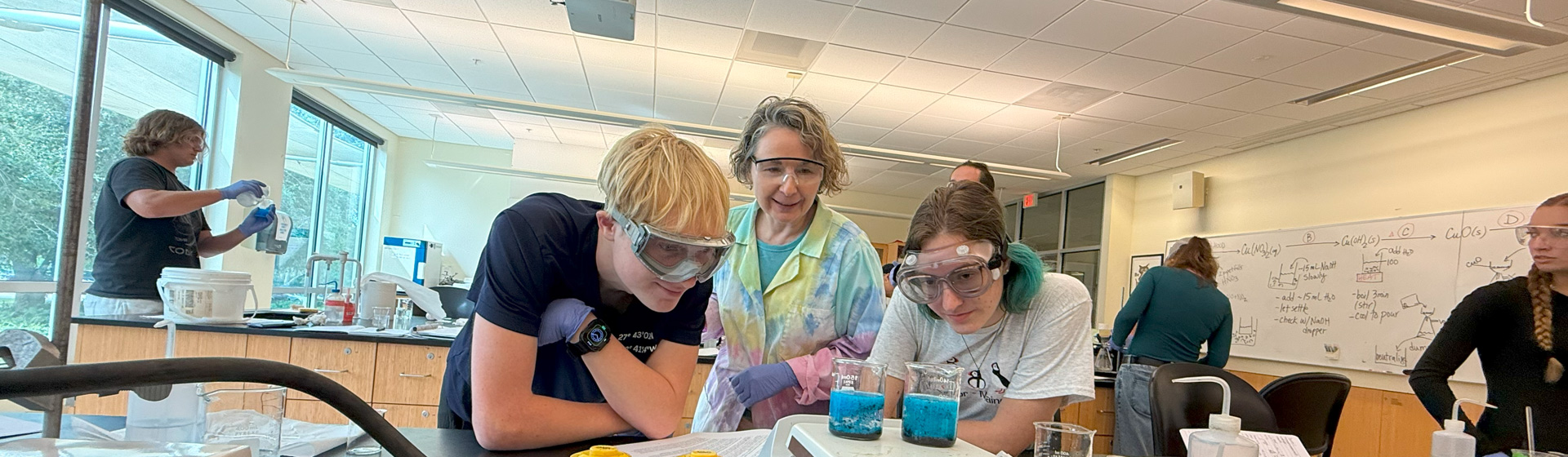 Two students wearing goggle conduct chemistry experiment in a lab while professor in tie-dye looks on