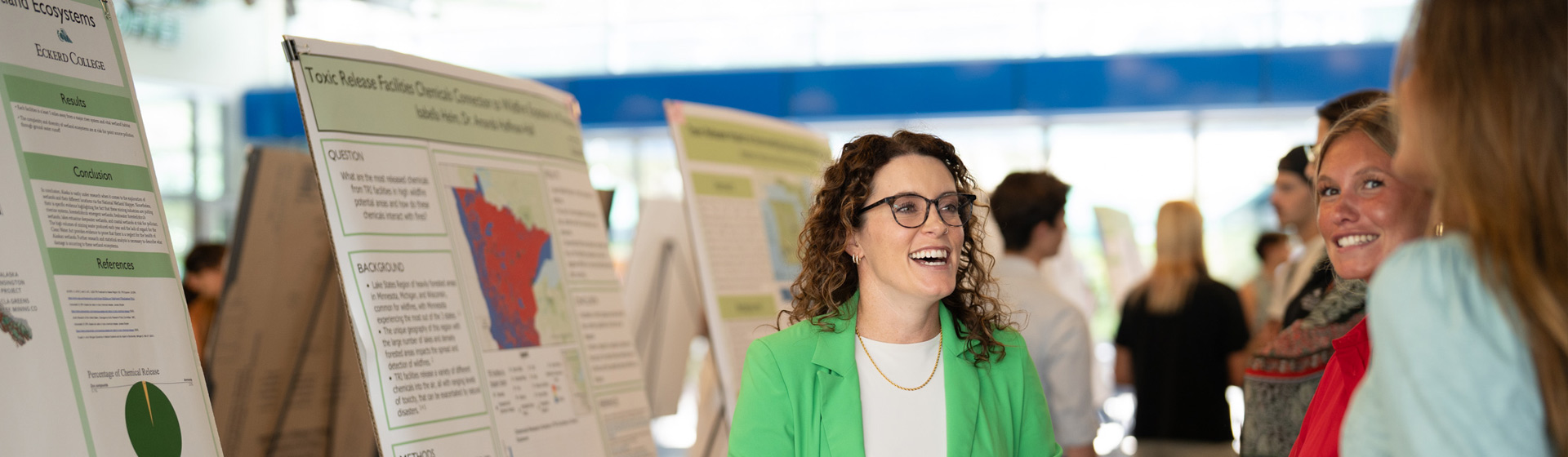 Professor wearing glasses laughs with two students who are presenting their scientific posters in a large room