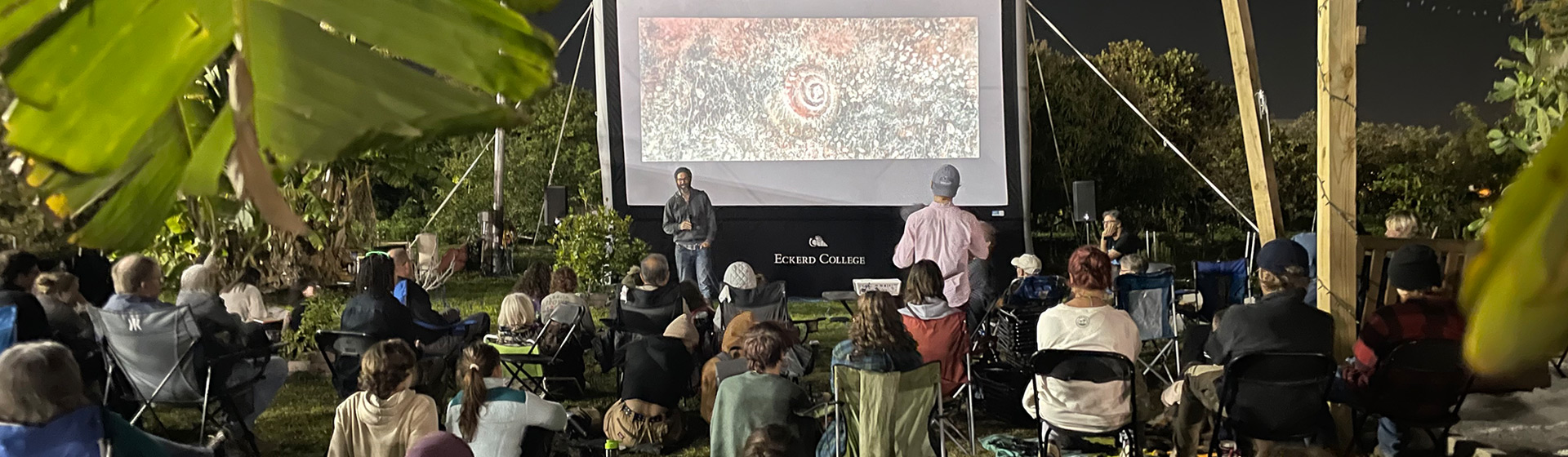 An audience seated outside in front of a large screen
