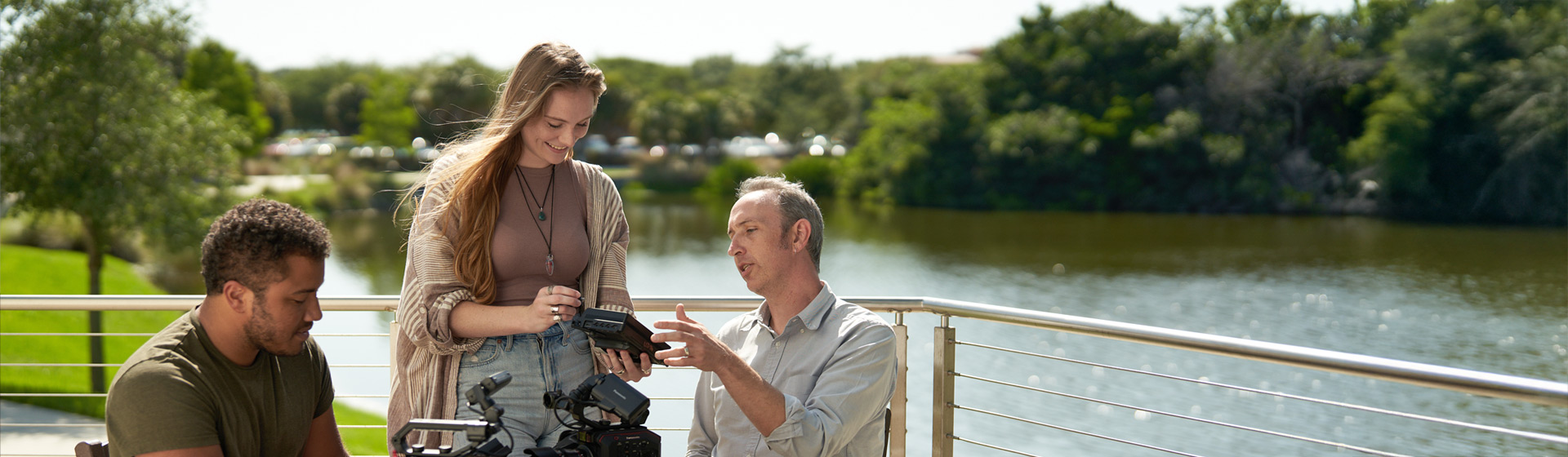 Professor shows camera equipment to two students seated outside by a pond