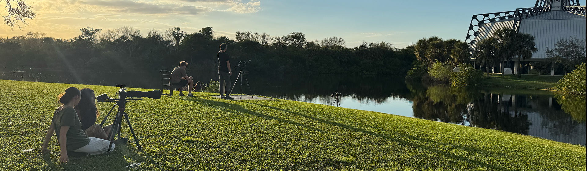 Students use a video camera on a tripod to capture outdoor scenery on a grassy slope by the water as the sun sets