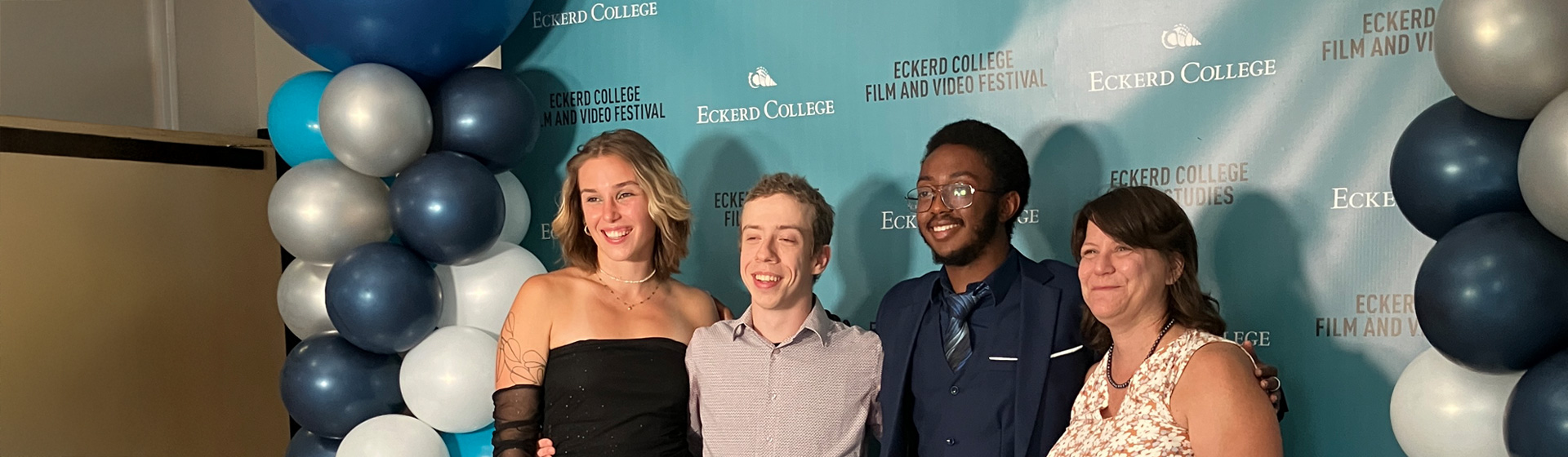 Students dressed in formal attire stand in front of blue and white balloons and a board that reads Eckerd College Film Studies