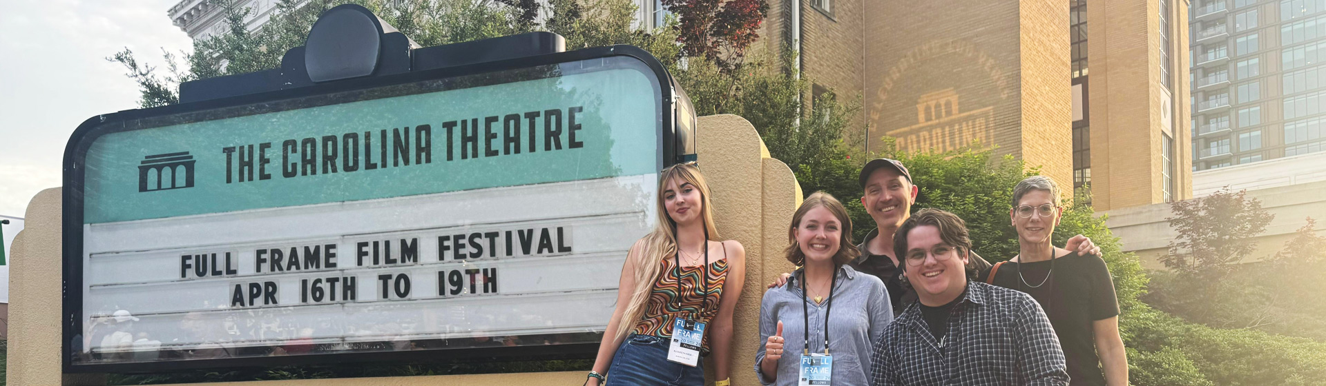 Small group of students and two professors stand by a sign outside a building that reads CAROLINA THEATRE FULL FRAME FESTIVAL