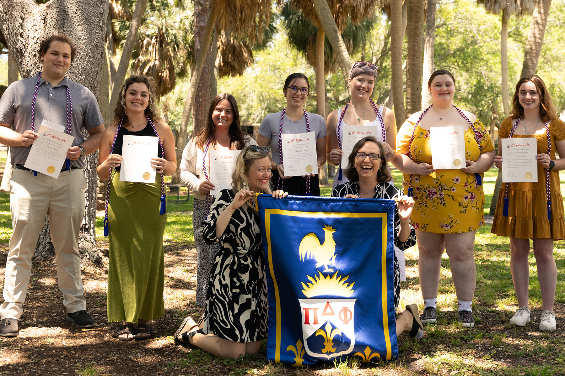 Seven students stand proudly with two faculty members holding up the honor society banner underneath the shade of palm trees