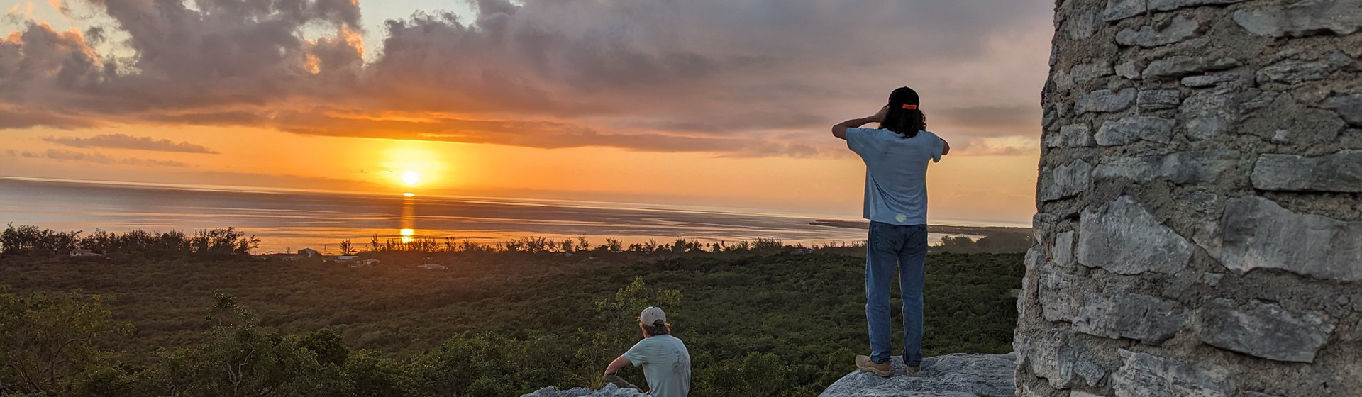 Two students stand on a rock structure looking at a sunset over the water using binoculars