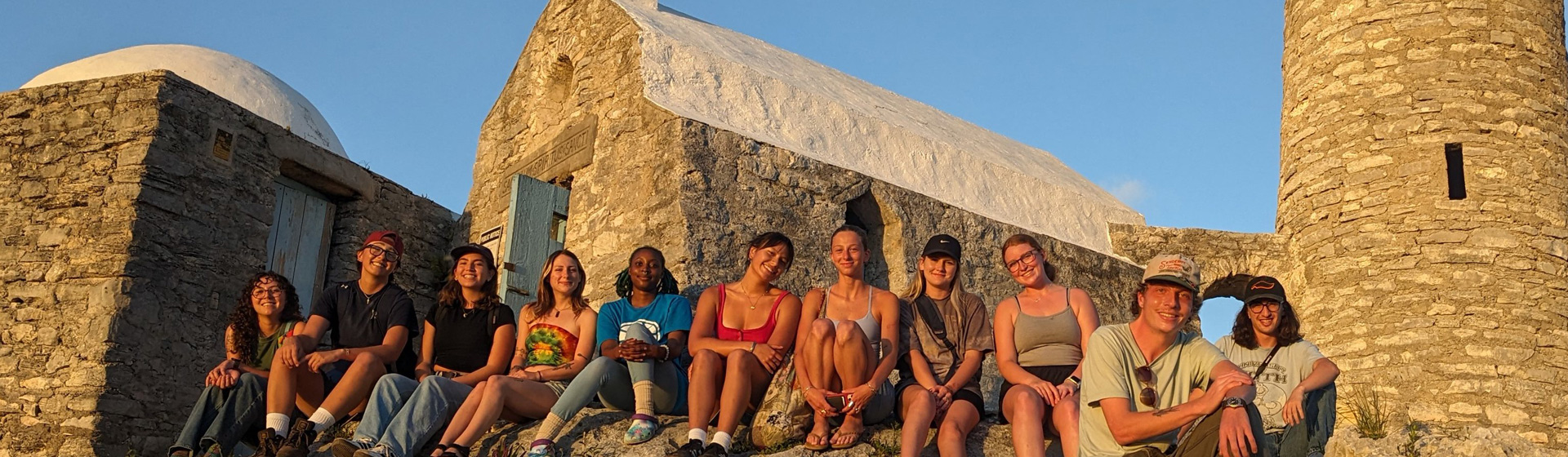 Nine college students seated in a row atop a concrete housing structure with a bright blue door bathed in sunlight from a sunset