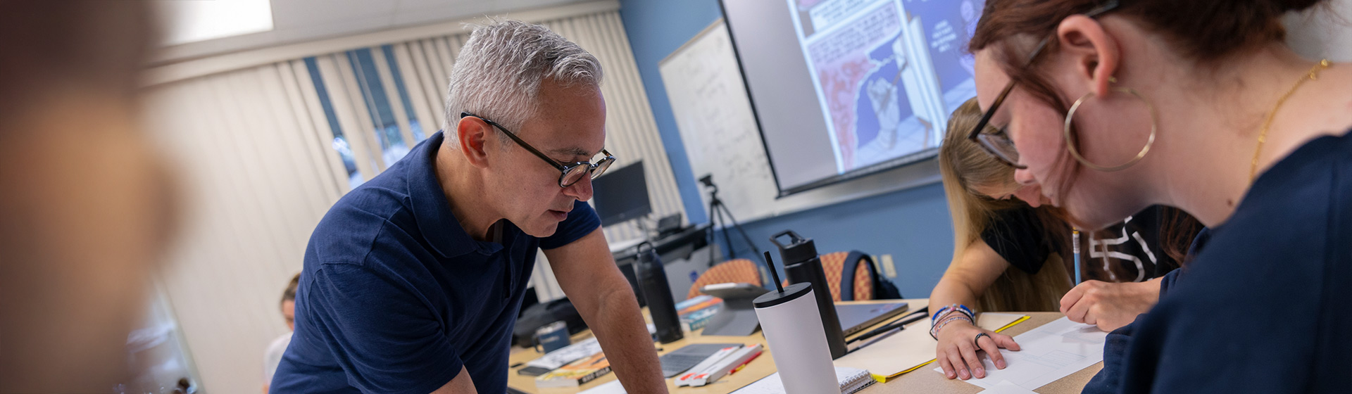 Professor in glasses leans over desk as student write on paper in front of a screen displaying a cartoon