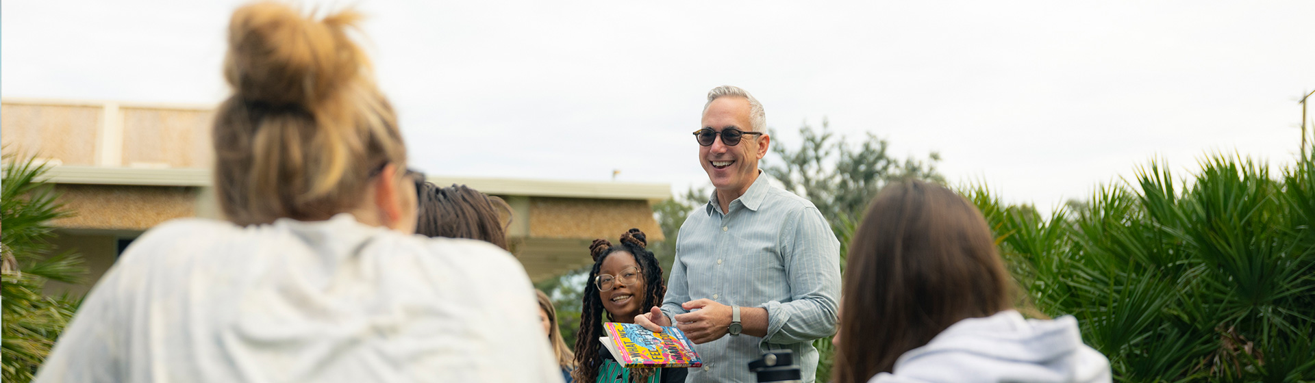Professor in glasses stands outside while talking to students seated around him and smiling
