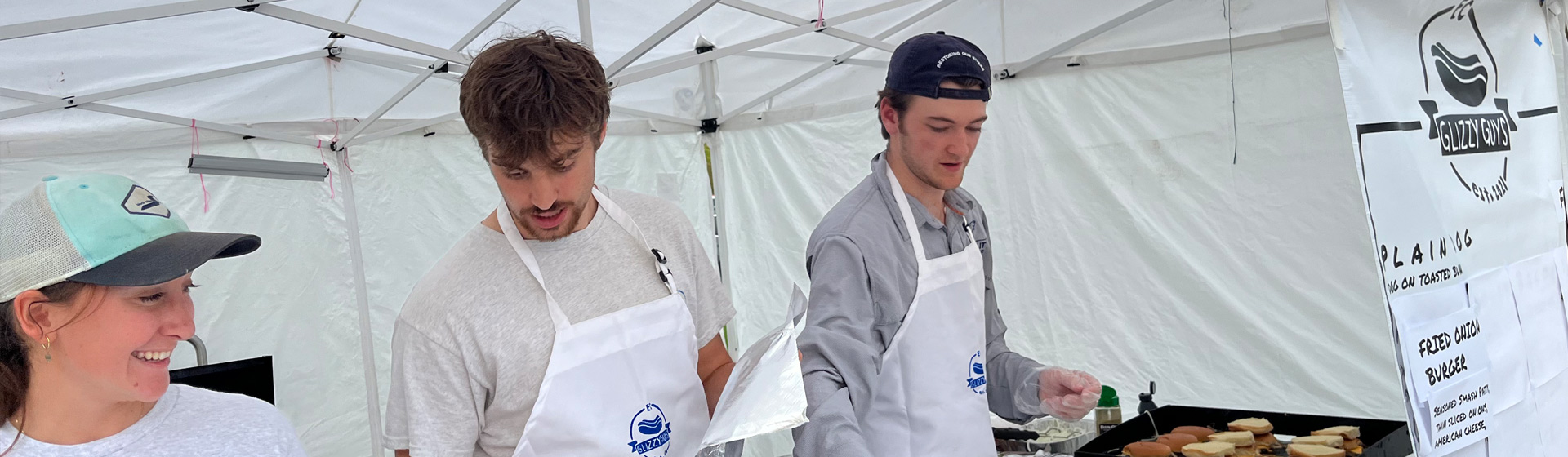 Three students wearing aprons and hats serve food next to a sign that lists prices for burgers
