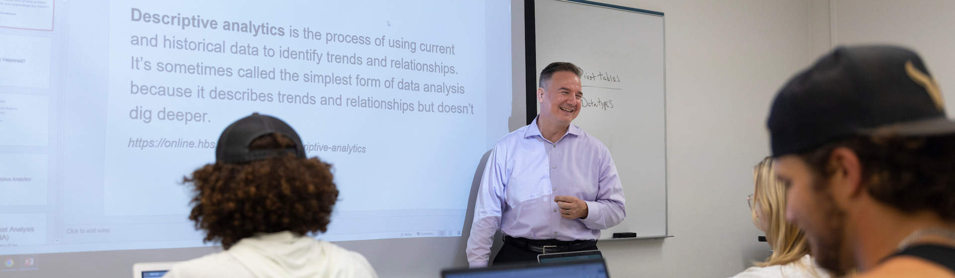 Professor stands before class next to screen with text about descriptive analyses