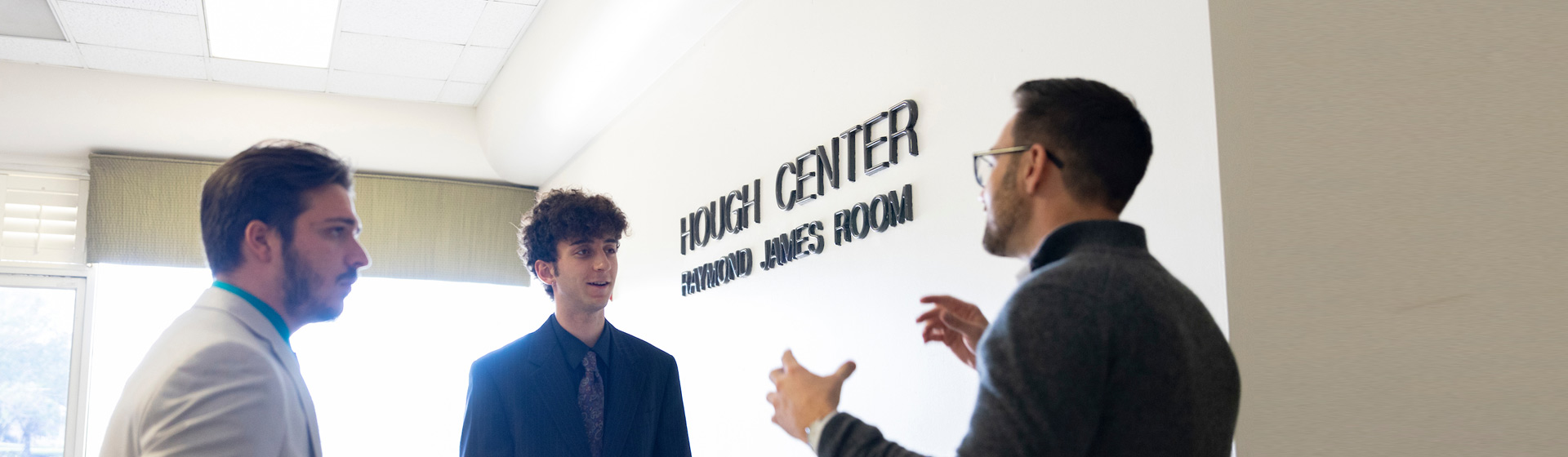 Two students in formal attire chat with staff person in room with Hough Center text on the wall