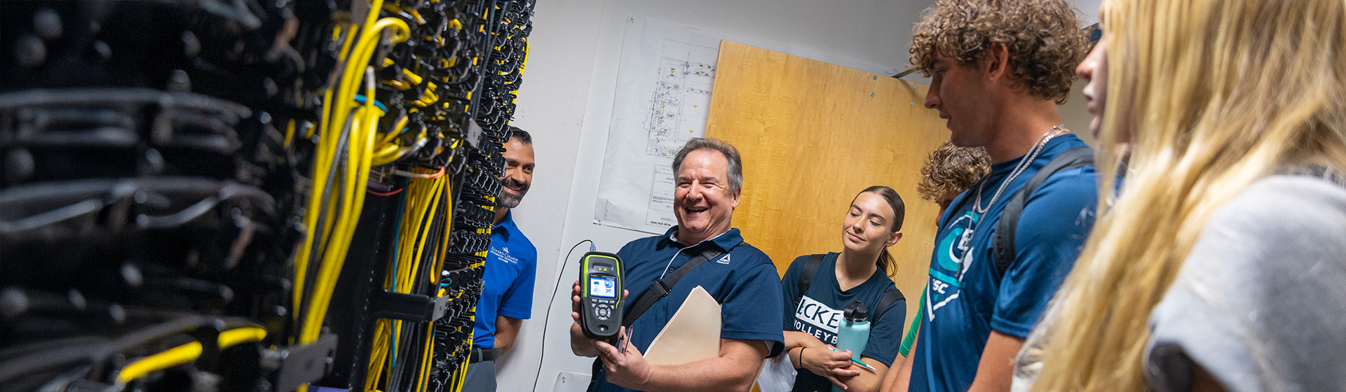 Students stand around a professor who shows them a tier of communication and network cables and a handheld monitoring device