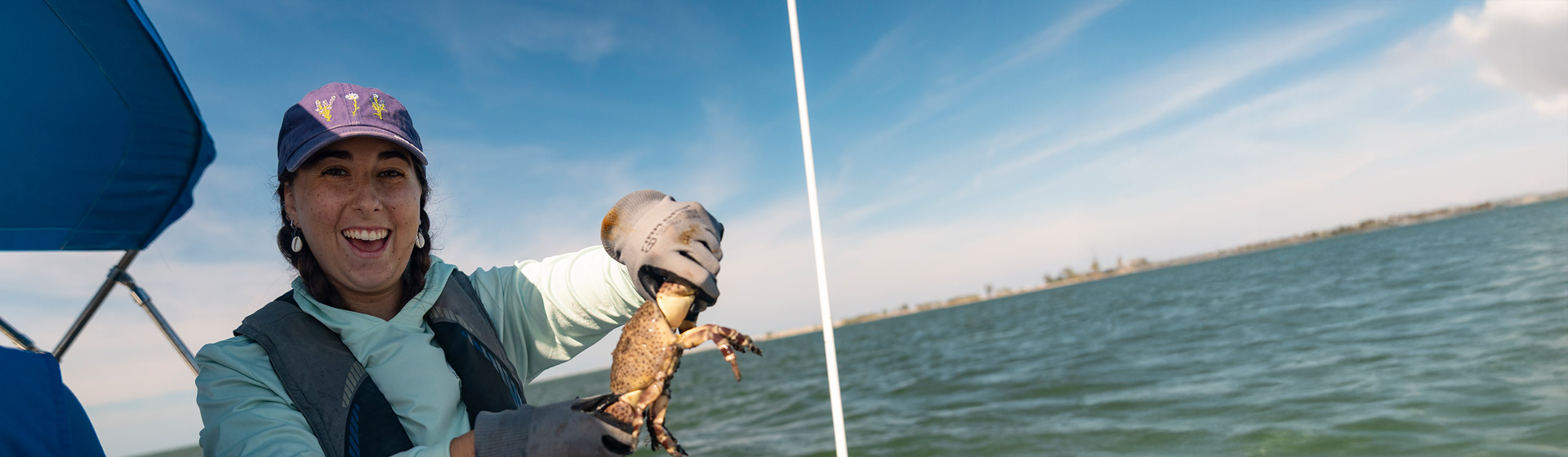 Student on boat wearing gloves holds up a big crab while smiling