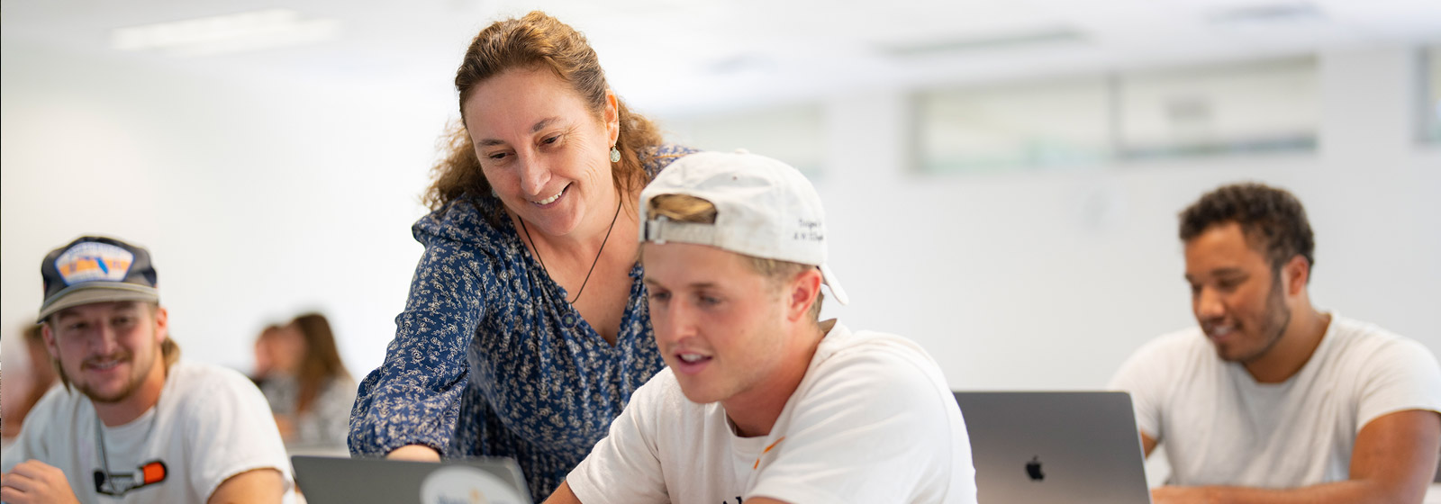 Professor leans over student wearing hat backwards to point at laptop while another male student in hat looks on and smiles