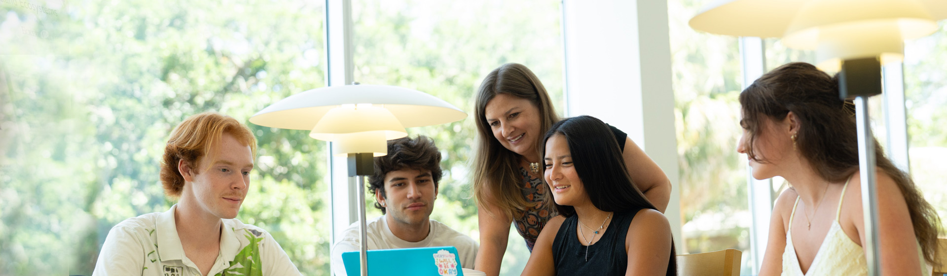 Professor leans over a group of four students to look at a laptop with them in a room by very tall windows with trees in the background