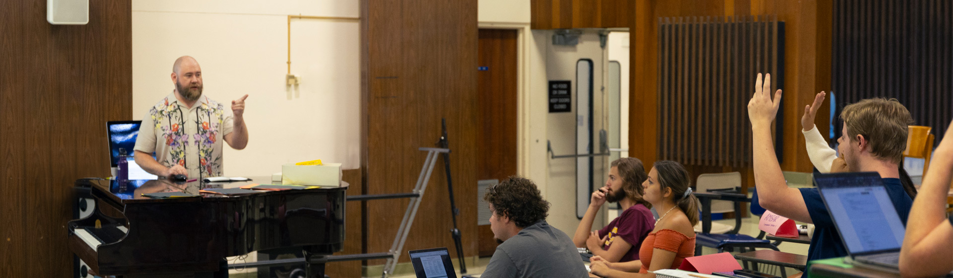 Professor standing at piano and pointing at students seated near him, who are raising their hands