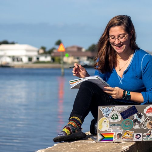 Student writing in notebook next to laptop on seawall