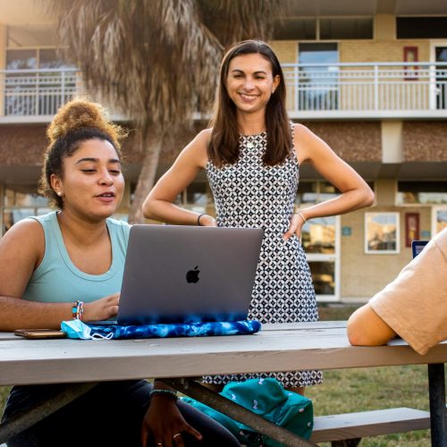 Professor stands with hands on hips and smiles as student works on laptop at outdoor table