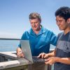 Professor and student stand on a dock while looking at a laptop