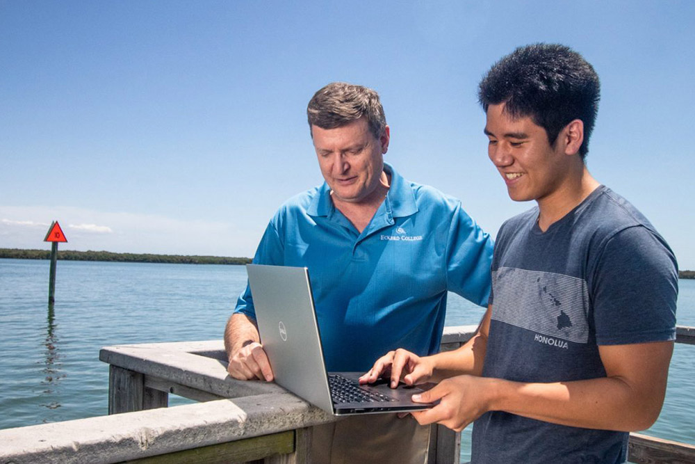 Professor and student stand on a dock while looking at a laptop