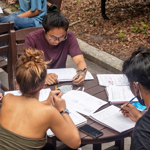 Students sit at a round table outside looking at notebooks together