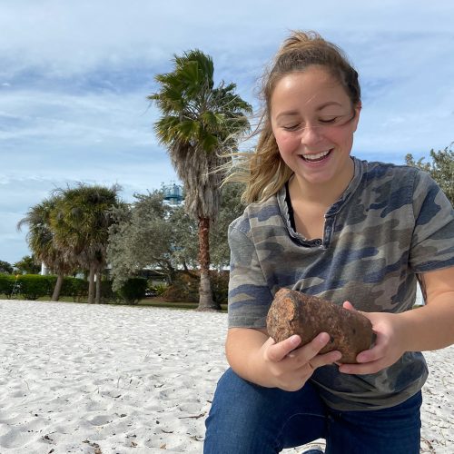 Student on one knee on the beach holding rock samlple