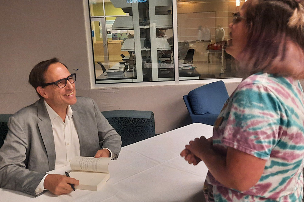 Author seated at table, signing book and talking to student