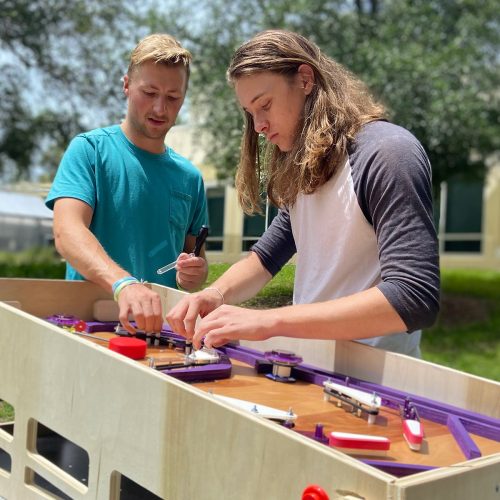 Two students working on a pinball machine outside