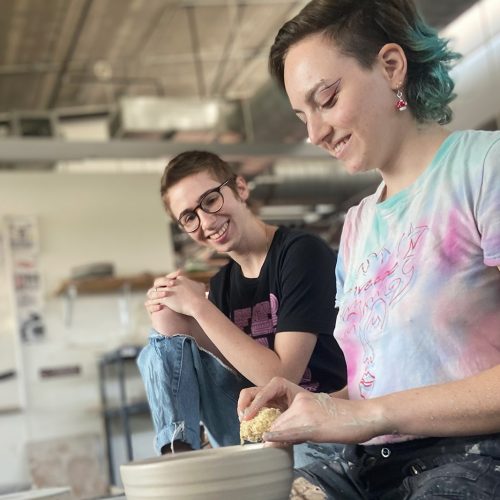 Student throwing pottery on the wheel while another looks on encouragingly