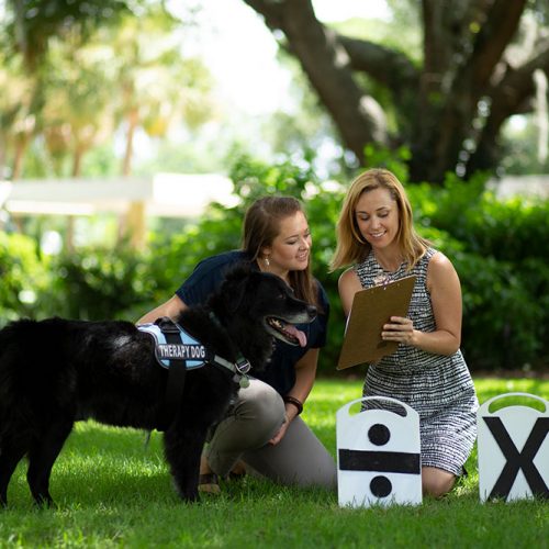 Dr. Lauren Highfill and an alumna knelling in the grass looking at a clipboard as a black dog stands at attention.