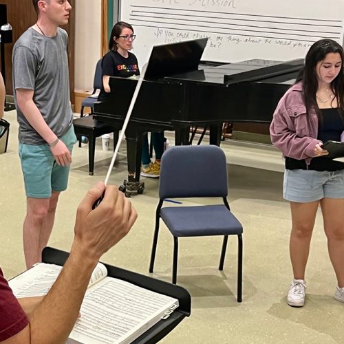 Two opera students receive instruction in a classroom as a pianist plays in the background.