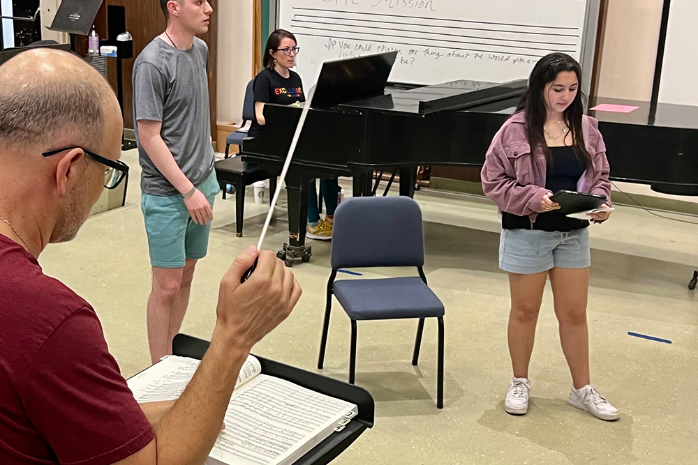 Two opera students receive instruction in a classroom as a pianist plays in the background.