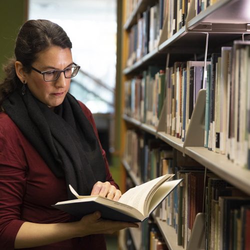 Professor wearing glasses, looking at a book in the library stacks