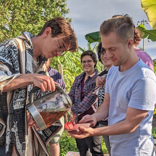 Student pouring tea into a vessel held by another student while standing in a garden