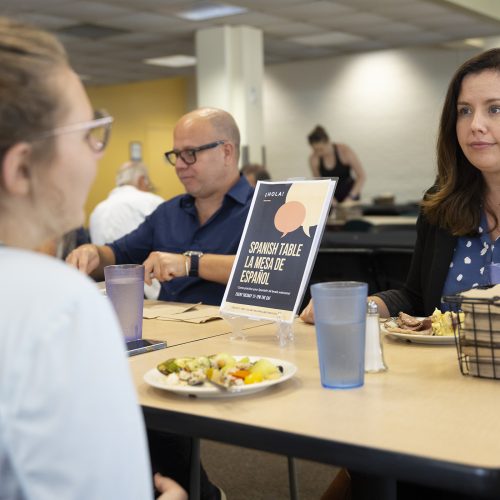 Cristina Delano speaking with a student while seated in the cafeteria.